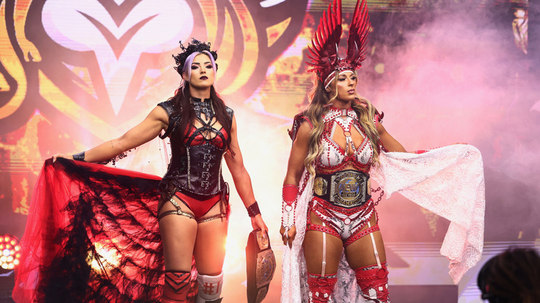 Divine Dominion, Lena Kross and Megan Bayne, pose on the stage, the AEW Women's World Tag Team Championships around their waists, before a match.