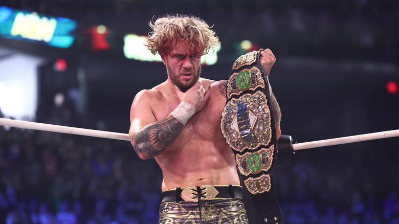 Will Ospreay holding his throat with his right hand and the AEW world title belt in his left hand