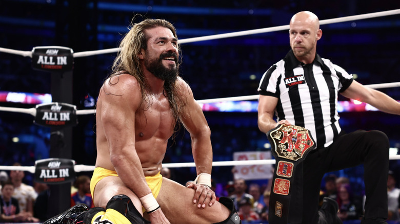 Andrade kneeling in the ring as the referee holds his National Championship belt behind him