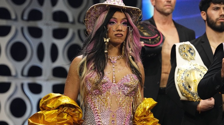 Mercedes Mone stands defiantly in a pink and gold outfit, with her many titles held by men in blazers.