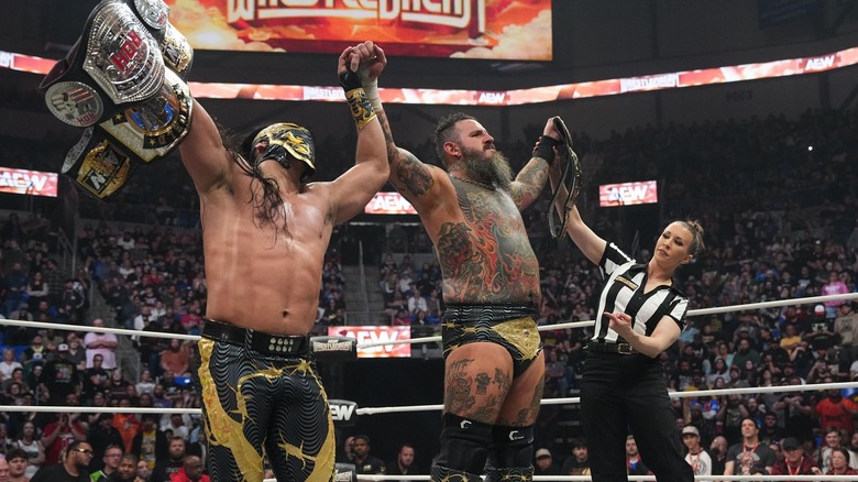 Bandido holds up his ROH World Championship and his AEW Tag Team Championship in one hand, and Brody King's hand in the other.