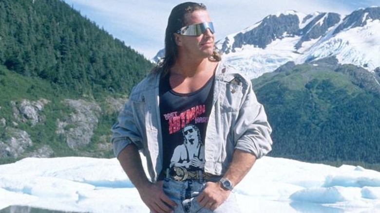 Bret Hart in double white denim, branded tank top and Hitman boots in front of a lake in Canada.