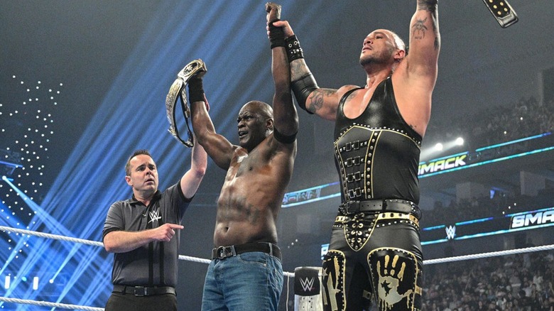A referee holds up the arms of Damian Priest and R-Truth as they celebrate a retention of their WWE Tag Team Championships on an episode of 