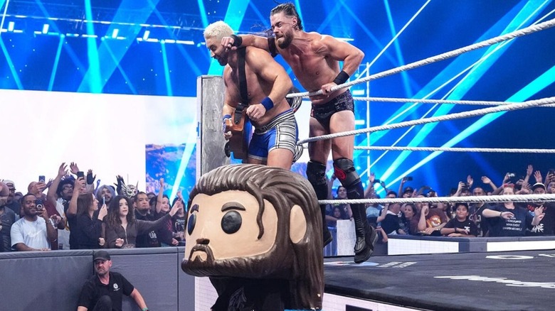 JD McDonagh hits Joe Hendry, who stands on the ring apron with a guitar, before a match, and a person wearing a costume Funko Pop head designed to look like McDonagh stands at ringside.
