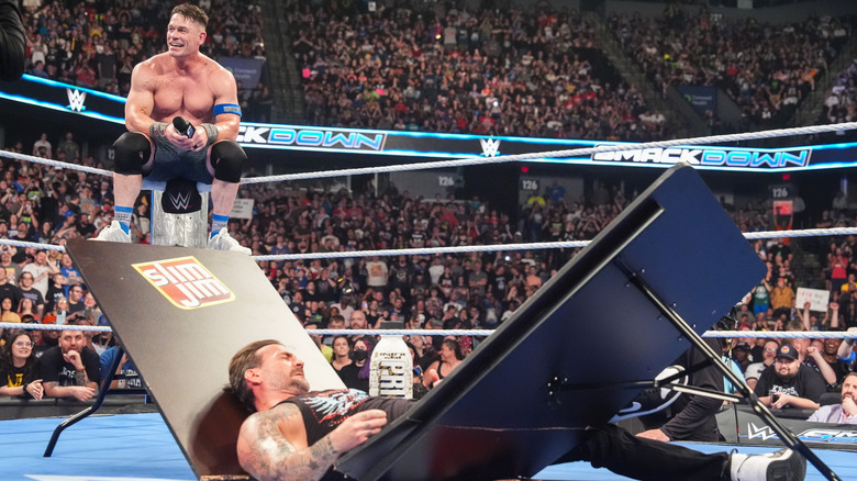 John Cena sitting on the turnbuckle with CM Punk in the ring lying through a broken table