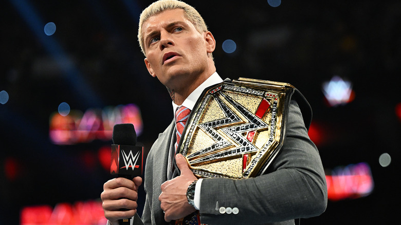 Cody Rhodes holds the Undisputed WWE Championship on his shoulder, and a microphone in the opposite hand.