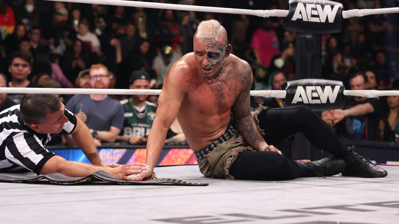 Darby Allin recovers in the ring, a referee and the AEW World Championship by his side, after a match on 
