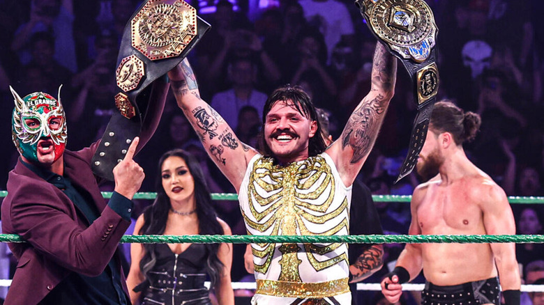Dominik Mysterio stands alongside El Grande Americano, Roxanne Perez, and JD McDonagh as he holds up both the AAA Mega Championship and the WWE Intercontinental Championship.