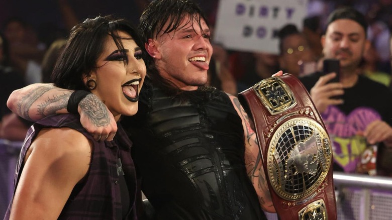 Dominik Mysterio, sticking out his tongue, stands on the ramp with Rhea Riple