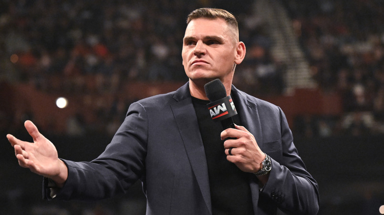 An image of WWE star Gunther wearing a sports coat, gesturing to his right while holding a micrphone