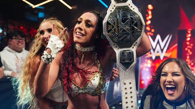 Jacy Jayne taunting with the NXT Women's Championship while Lainey Reid and Fallon Henley laugh beside her.