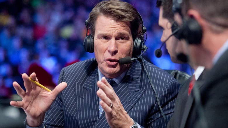 John Bradshaw Layfield sits at the commentary desk and motions toward Michael Cole during an episode of WWE programming.