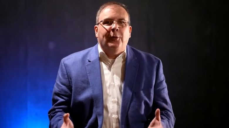 Kevin Kelly speaking in front of a black and blue background wearing a suit