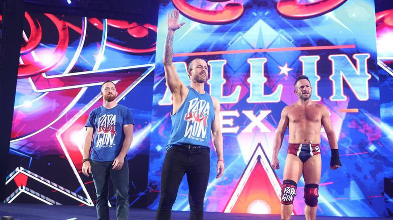 Adam Cole waves to fans from the stage at All In, flanked by Kyle O'Reilly and Roderick Strong after relinquishing his TNT Championship.