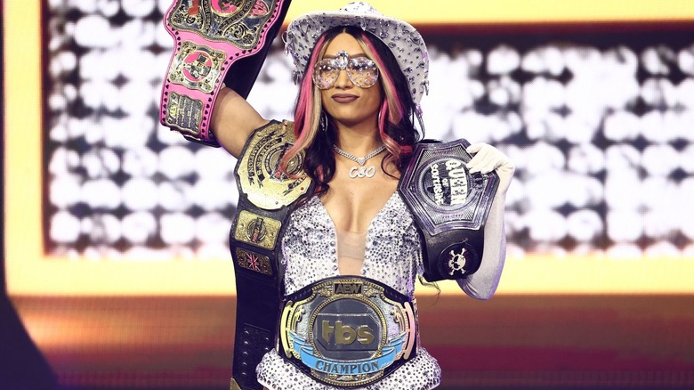Mercedes Moné walks in with the Owen Hart cup title hoisted in the air, and her two RevPro titles and AEW TBS title hanging off her body.