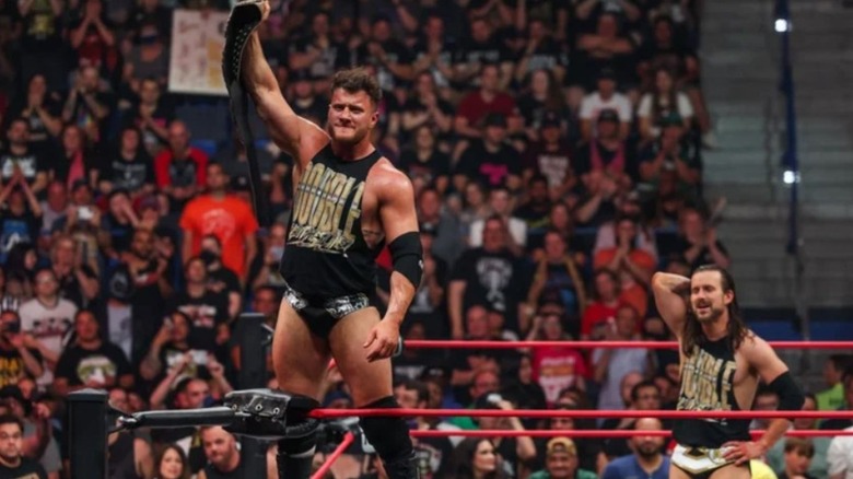 MJF holds up the AEW World Championship while standing on the turnbuckle as Adam Cole looks on in the ring during an episode of AEW programming.