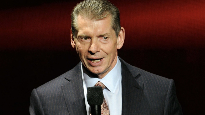 Vince McMahon speaks into a microphone in front of a dark red and black screen.