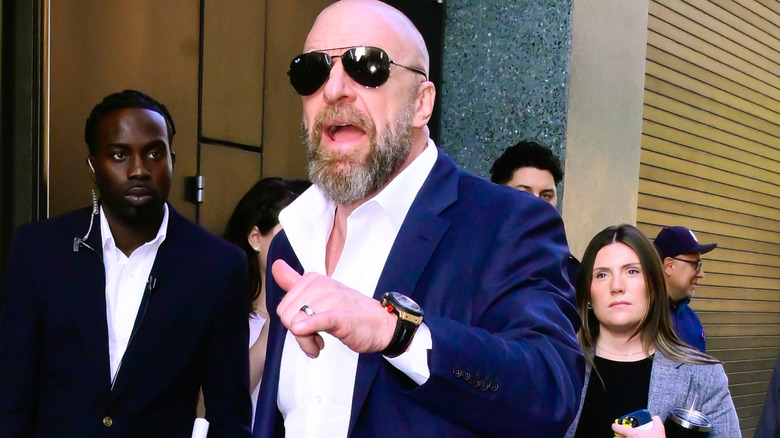 Triple H holds his hand out, with his thumb and pinky sticking out, and speaks to an off-screen recepient.