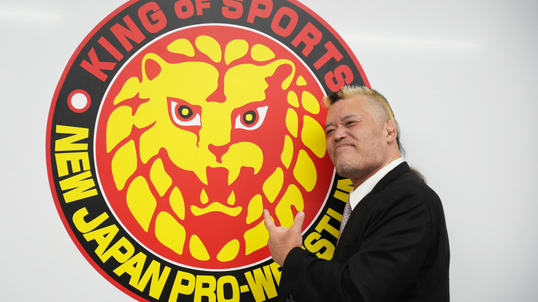 NJPW legend Hiroyoshi Tenzan stands in front of the company's emblem and points at it during a special press conference event
