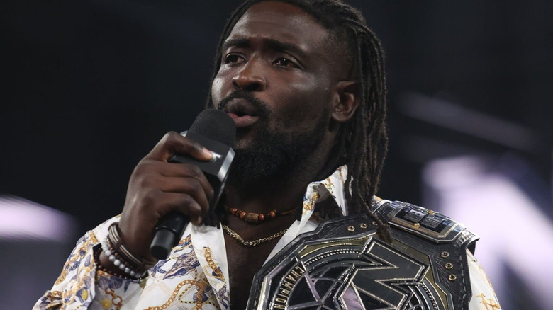 Oba Femi speaks on the microphone with his NXT Championship on his shoulder.