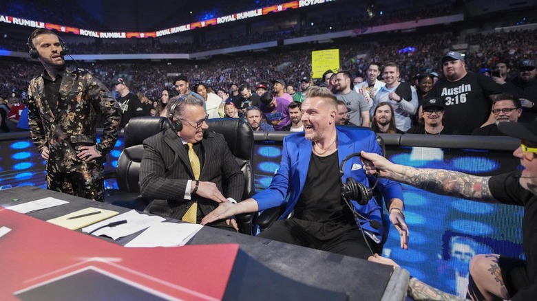 Pat McAfee shakes Michael Cole's hand as Corey Graves looks on in disgust.