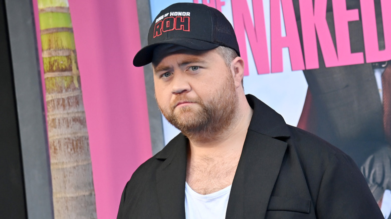 Paul Walter Hauser wearing a Ring of Honor hat in front of a poster for The Naked Gun