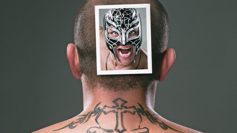 Rey Mysterio reveals the back of his head unmasked with a polaroid photo attached of what the front of his face looks like in a traditionally worn luchador styled mask