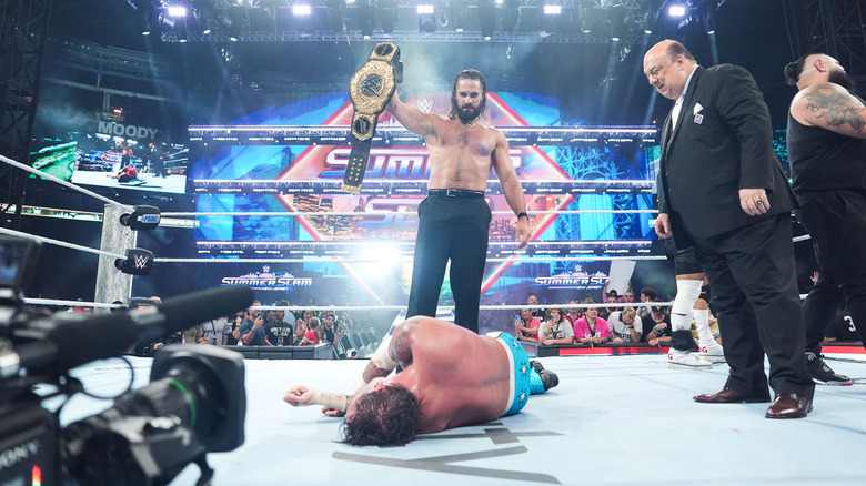 Seth Rollins holding up the WWE World Heavyweight Championship beside Paul Heyman and Bronson Reed over a laid-out CM Punk