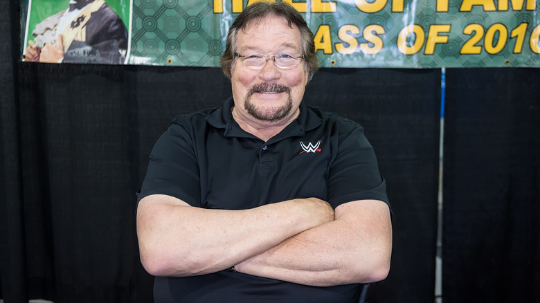 Ted Dibiase with his arms crossed smiling in a WWE polo shirt