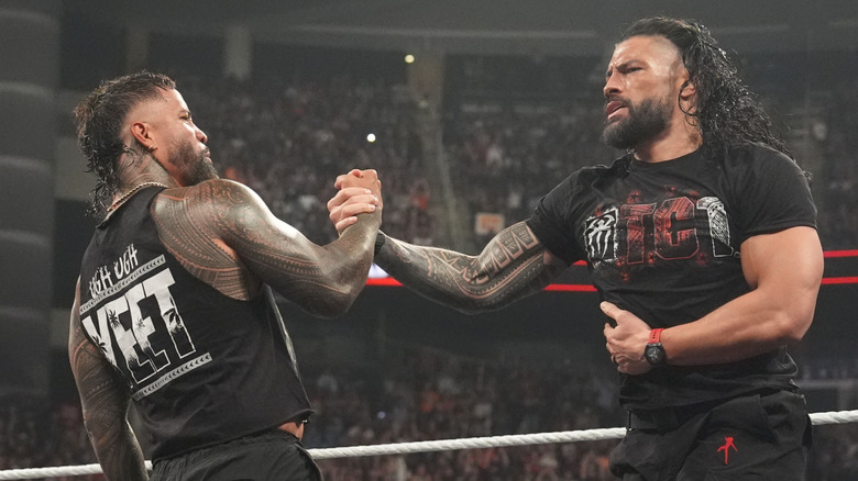 Roman Reigns and Jey Uso solidly shake hands as Reigns holds his side.