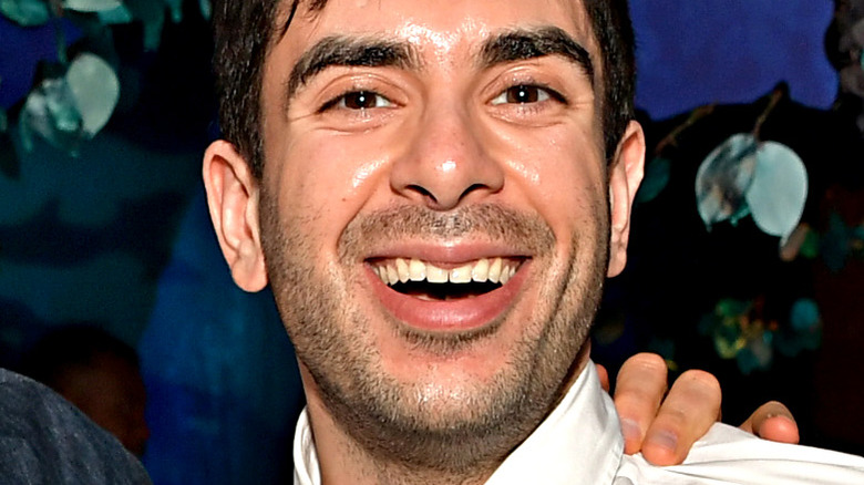 Khan smiling in front of blue background with hand on shoulder.