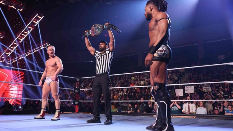 A referee stands between Ilja Dragunov and Trick Williams, holding up the NXT Championship, before the pair's match at Vengeance Day 2024.