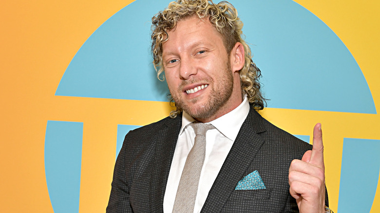 Kenny Omega pointing upward while wearing a suit