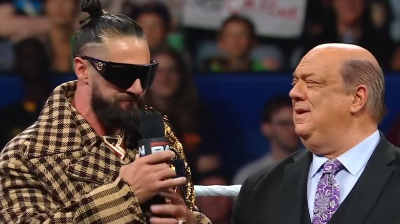 Seth Rollins speaks into a WWE microphone while Paul Heyman looks on