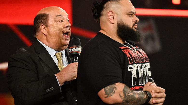 Paul Heyman speaks on the microphone behind an imposing Bronson Reed.