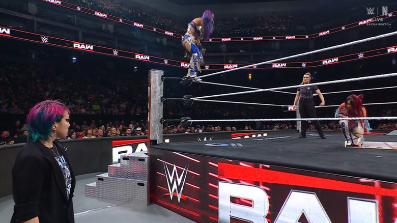 Asuka watching from the outside as Sane ascends to the top rope and SKY is inside the ring