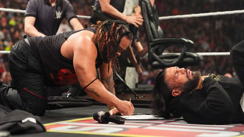 Jacob Fatu signs the contract in the ring, as World Heavyweight Champion Roman Reigns grabs his throat, feeling the impact of the Tongan Death Grip, during an episode of 