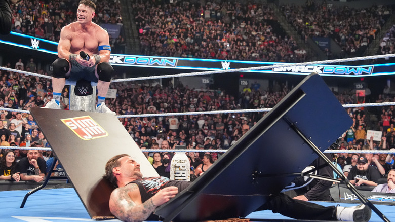 John Cena sits on a ring post addressing a fallen CM Punk, lying on a broken table, on "WWE SmackDown"