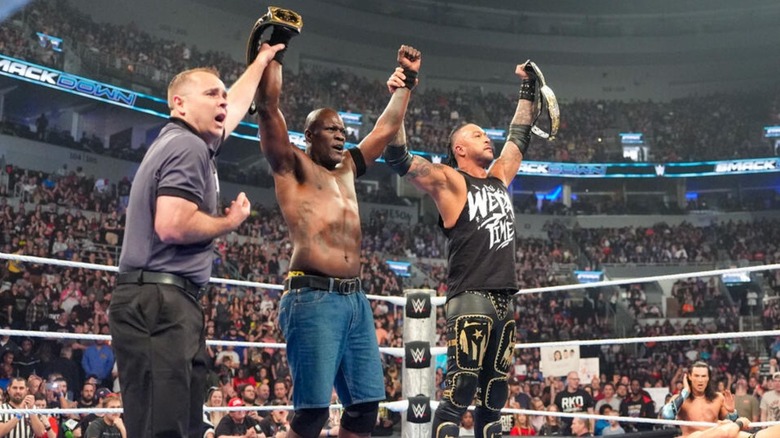 A referee holds up the arm of R-Truth as he grasps Damian Priest after a successful retention of the WWE Tag Team Championships.