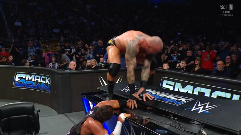 Orton about to stomp on Cardona's arm on top of the announce desk