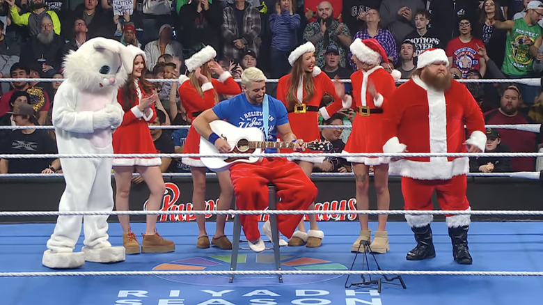 Joe Hendry is surrounded by several dressed up figures (one Easter Bunny, five Santas, and one Elf on the top rope).