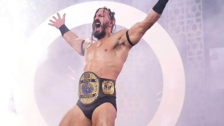 Pac poses on stage, AEW World Trios Championship around his waist, before a match on AEW programming.