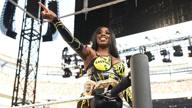 Naomi smiles widely as she stands on the second rope, WWE Women's World Championship around her waist and finger pointed towards an off-screen subject.