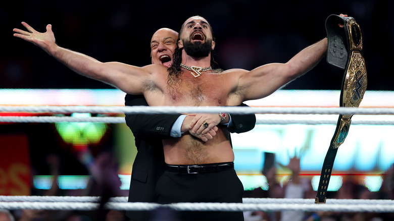 Paul heyman hugs Seth Rollins from behind as he holds the WWE World Heavyweight Championship.