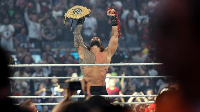 Roman Reigns holds up his title belt and the ula fala