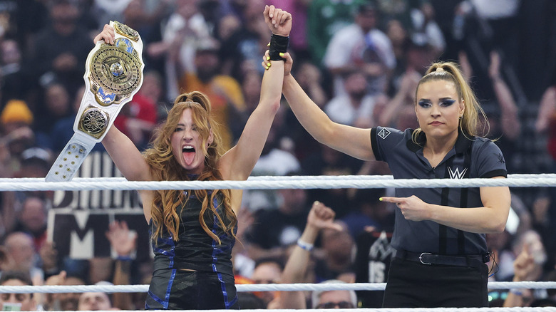 Referee Jessika Karr holds up Becky Lynch's hand as she hoists up her third WWE Women's Intercontinental Championship, tongue out.