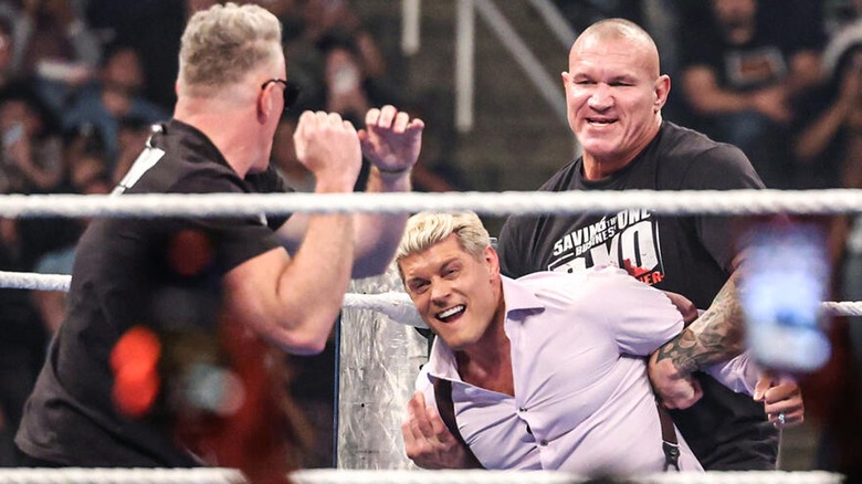 Pat McAfee preparing to slug Cody Rhodes with a punch while Randy Orton subdues him.