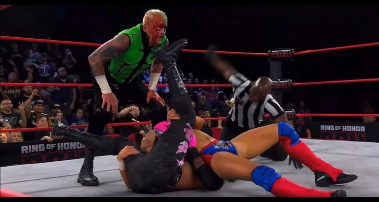 Dustin Rhodes in Green Lantern-inspired outfit while Sammy Guevara pins someone wearing red and blue Superman gear